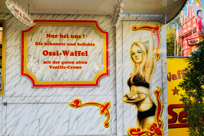 Airbrush an einem Wagen auf der Kirmes, der Waffeln verkauft. Zu sehen ist ein blondes Mädchen im Bikini mit einem Tablett mit einer Waffel. Im Text daneben steht "Ossiwaffel".