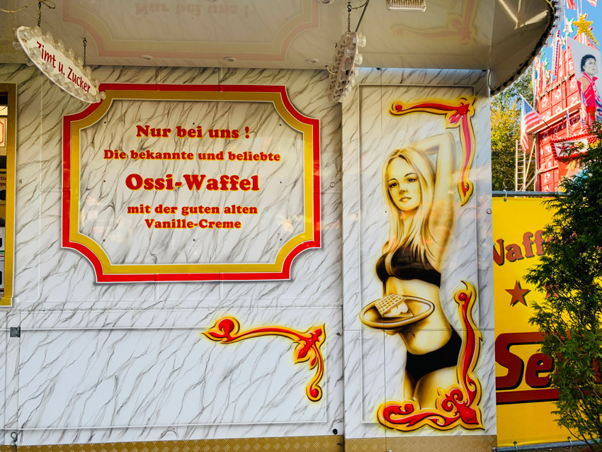 Airbrush an einem Wagen auf der Kirmes, der Waffeln verkauft. Zu sehen ist ein blondes Mädchen im Bikini mit einem Tablett mit einer Waffel. Im Text daneben steht "Ossiwaffel".