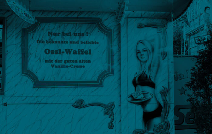 Airbrush an einem Wagen auf der Kirmes, der Waffeln verkauft. Zu sehen ist ein blondes Mädchen im Bikini mit einem Tablett mit einer Waffel. Im Text daneben steht "Ossiwaffel".
