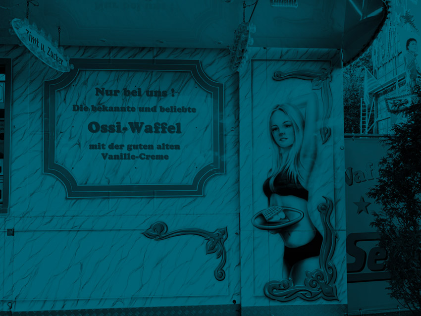 Airbrush an einem Wagen auf der Kirmes, der Waffeln verkauft. Zu sehen ist ein blondes Mädchen im Bikini mit einem Tablett mit einer Waffel. Im Text daneben steht "Ossiwaffel".