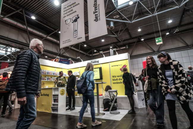 Der Messestand von "der Freitag" auf der Leipziger Buchmesse 2023. Der Stand ist gelb gestaltet, einige Personen stehen darauf und davor.