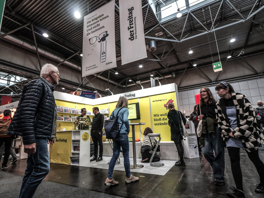 Der Messestand von "der Freitag" auf der Leipziger Buchmesse 2023. Der Stand ist gelb gestaltet, einige Personen stehen darauf und davor.